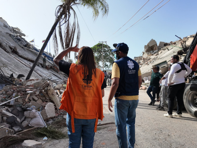 Caritas-Helfer*innen in Venezuela gehen zwischen Trümmern eingestürzter Gebäude nach einem Erdbeben eine Straße entlang, im Hintergrund weitere Menschen und ein Einsatzfahrzeug.