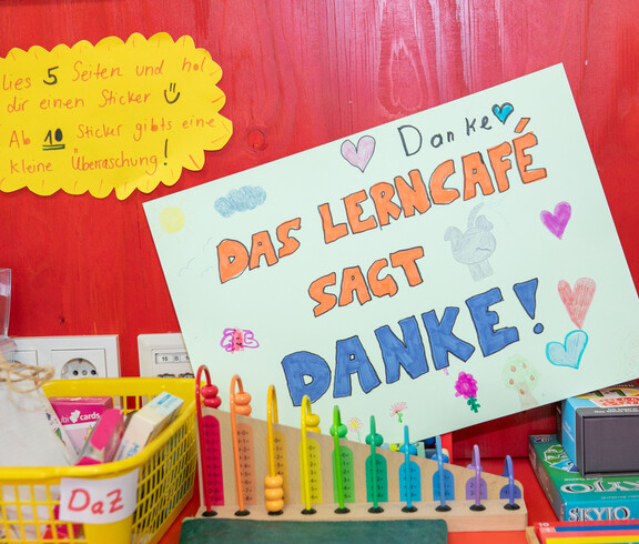 Selbstgestaltetes Plakat mit „Das Lerncafé sagt Danke!“ zwischen Lernmaterialien und Spielen an roter Wand.