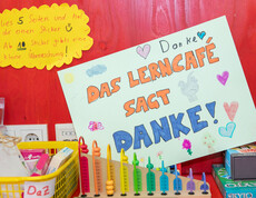 Selbstgestaltetes Plakat mit „Das Lerncafé sagt Danke!“ zwischen Lernmaterialien und Spielen an roter Wand.