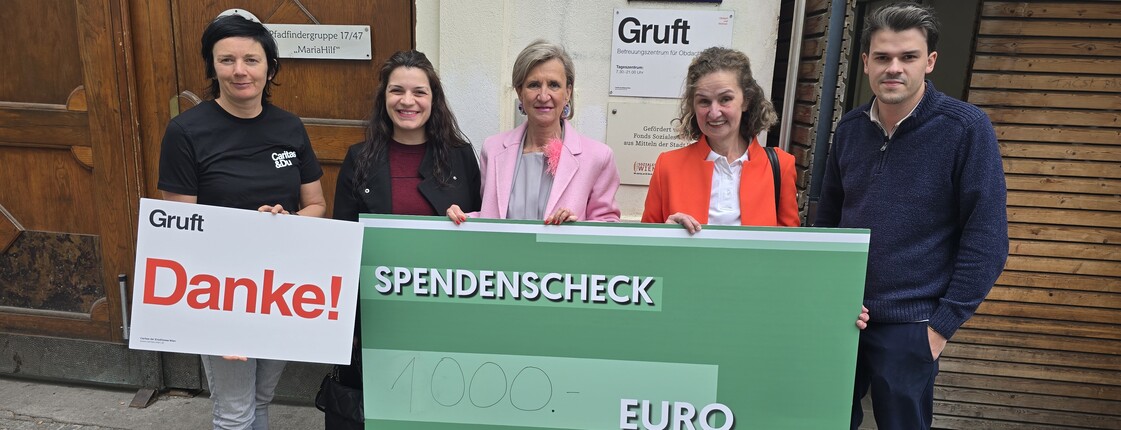 Fünf Personen stehen vor einem Gebäude und halten gemeinsam ein großes grünes Spendenscheck‑Plakat mit der Aufschrift „Danke! Spendenscheck 1.000 Euro“.