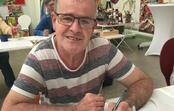 Künstler mit Brille malt mit Stift ein farbiges Bild im Atelier der Caritas-Kunstgruppe im Weinviertel.