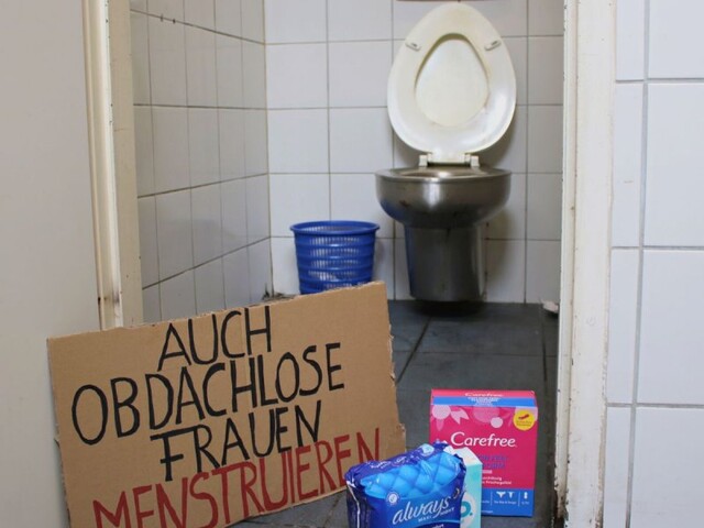  Ein handgeschriebenes Pappschild mit der Aufschrift „AUCH OBDACHLOSE FRAUEN MENSTRUIEREN“ steht neben Menstruationsprodukten vor einer geöffneten Toilettentür.