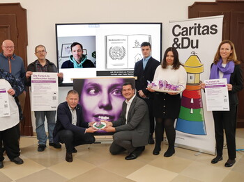 Gruppenfoto von neun Personen bei einer Veranstaltung der Caritas. Zwei Männer knien vorn und halten gemeinsam ein lilafarbenes Herzsymbol. Weitere Personen stehen dahinter, drei von ihnen halten Plakate mit der Aufschrift „Die Farbe Lila aufleuchten lassen.“ Eine Frau trägt einen Tablett mit lilafarben verpackten Geschenken. Im Hintergrund ist ein Bildschirm mit einem Videocall und eine Illustration zur Menschenrechtskonvention zu sehen. Rechts steht ein Roll-up mit der Aufschrift „Caritas & Du“ und einem bunten Leuchtturm.