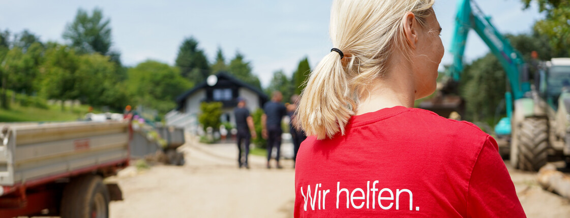 Eine Caritas Mitarbeiterin in einem roten Caritas T-Shirt mit der Aufschrift „Wir helfen“ steht auf einer unbefestigten Straße, während im Hintergrund Bauarbeiten und Menschen vor einem Haus zu sehen sind.