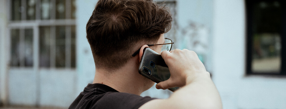 Ein junger Mann telefoniert mit dem Handy auf einer Bank vor einem weißen Haus.