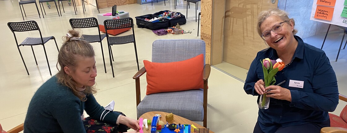 Mehrere Personen sitzen an einem Tisch und arbeiten gemeinsam mit bunten Bausteinen und Materialien in einem Innenraum.