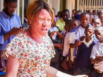 Eine Frau in einem geblümten Kleid spricht gestikulierend zu einer großen Gruppe von Schüler*innen, die eng beisammenstehen und aufmerksam zuhören; im Hintergrund stehen weitere Erwachsene und Jugendliche im Schulflur.