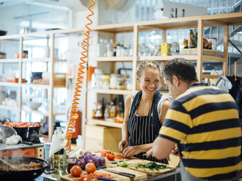 Zwei Personen bereiten gemeinsam frisches Gemüse in einer offenen Gemeinschaftsküche zu. Beide tragen Schürzen, schneiden Zutaten und lächeln einander an. Im Hintergrund sind Regale mit Kochutensilien und Lebensmitteln zu sehen. 
