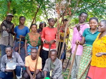 Eine Gruppe von Frauen und Männern steht gemeinsam mit Werkzeugen in einer bewaldeten ländlichen Umgebung.