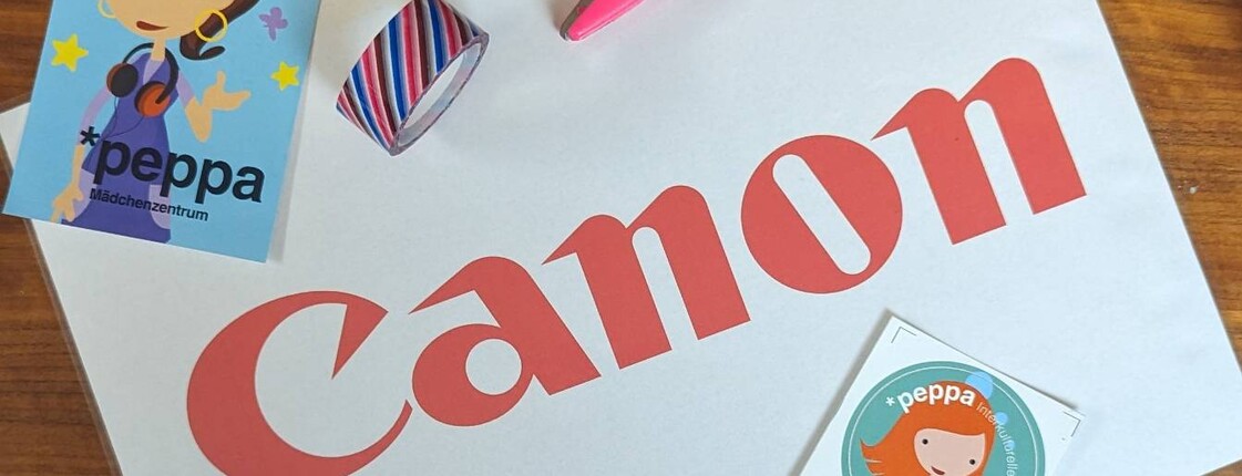Foto einer Arbeitsfläche aus Holz mit einer Canon‑Kartonverpackung, Bastelmaterialien wie Schere, Zange, Klebestift, Faden und bunten Stickern mit einer gezeichneten Figur.