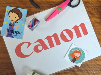 Foto einer Arbeitsfläche aus Holz mit einer Canon‑Kartonverpackung, Bastelmaterialien wie Schere, Zange, Klebestift, Faden und bunten Stickern mit einer gezeichneten Figur.