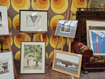 Präsentation gerahmter zeitgenössischer Kunstwerke auf Staffeleien und am Boden in einem Ausstellungsraum mit gemusterter Tapete.