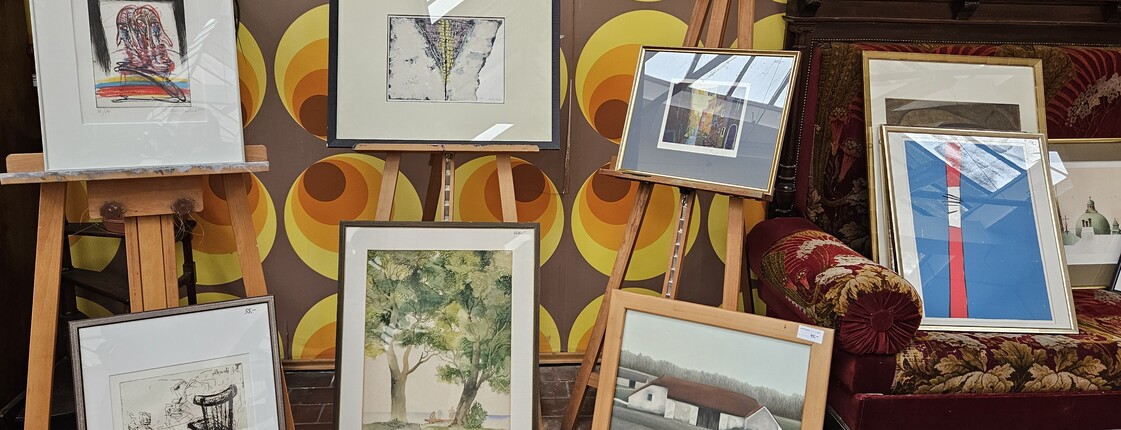 Präsentation gerahmter zeitgenössischer Kunstwerke auf Staffeleien und am Boden in einem Ausstellungsraum mit gemusterter Tapete.