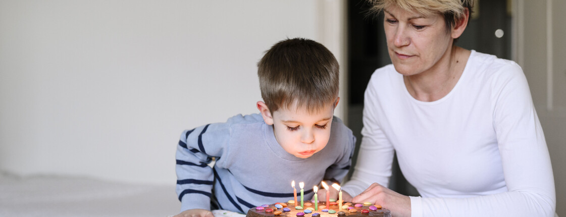 Kind pustet Kerzen auf Schokoladenkuchen aus, erwachsene Person sitzt daneben am Tisch in einem Mutter-Kind-Haus