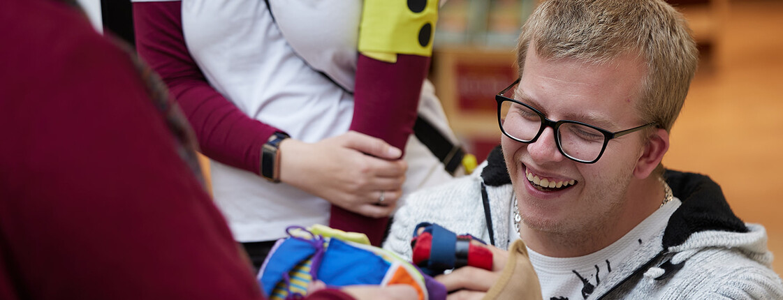 Person mit Brille probiert bei einer Caritas-Veranstaltung Hilfsmittel aus, die verschiedene Einschränkungen erlebbar machen. Die Person trägt einen gepolsterten Handschuh und erkundet gemeinsam mit anderen Teilnehmenden einen bunten Stoffgegenstand. Im Hintergrund ist ein T-Shirt mit dem Aufdruck „Caritas & Du“ zu sehen.