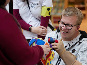 Person mit Brille probiert bei einer Caritas-Veranstaltung Hilfsmittel aus, die verschiedene Einschränkungen erlebbar machen. Die Person trägt einen gepolsterten Handschuh und erkundet gemeinsam mit anderen Teilnehmenden einen bunten Stoffgegenstand. Im Hintergrund ist ein T-Shirt mit dem Aufdruck „Caritas & Du“ zu sehen.