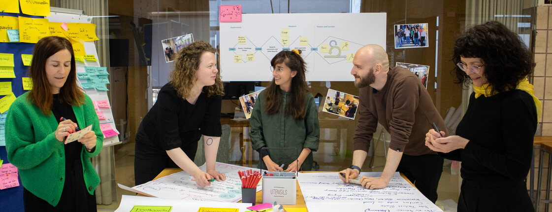 Gruppe von mehreren Personen steht um einen Tisch und arbeitet mit Notizen und Stiften an einem gemeinsamen Projekt