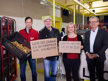 Vier Personen stehen in einer Lebensmittelausgabestelle der Caritas. Eine Person hält eine Kiste mit Kartoffeln, zwei andere halten Kartonschilder mit der Aufschrift „Wenn der Einkauf zur Existenzfrage wird: Wir helfen!“ und „Caritas-Hotline 05 17 76 300“. Im Hintergrund sind Regale und Lebensmittel zu sehen. Die Szene vermittelt Solidarität und Hilfe für Menschen in Not.