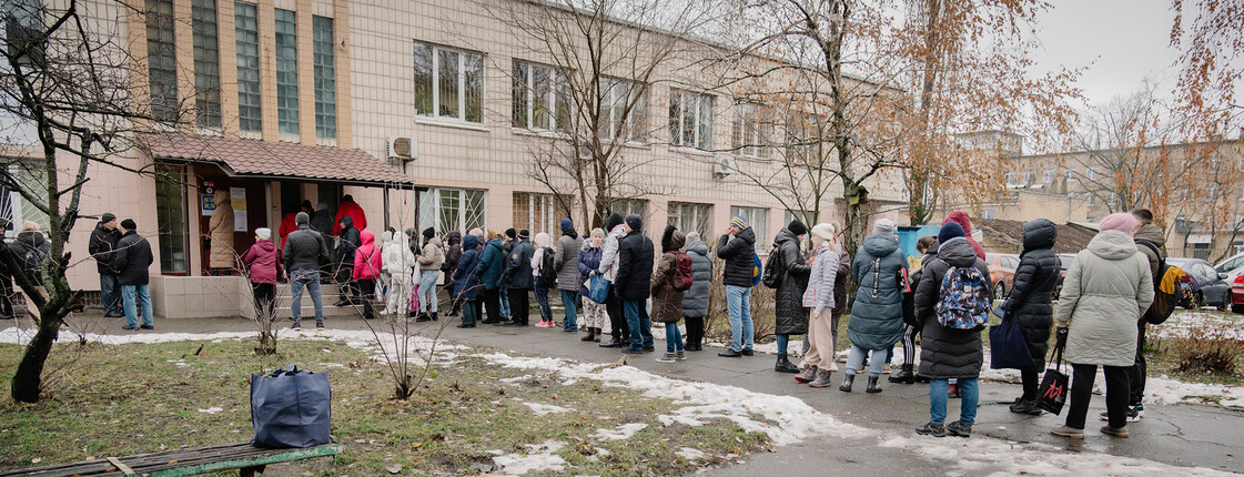 Ein Gebäudes, vor dem eine lange Schlange von Menschen im Winter ansteht und wartet. Im Vordergrund sind Pakete mit dem Aufdruck der Caritas und "Nachbar in Not" zu sehen.