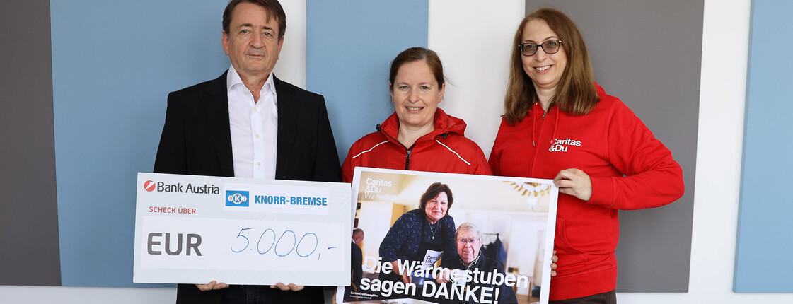 Drei Personen stehen vor einer Wand und präsentieren einen symbolischen Scheck über 5.000 Euro sowie ein Plakat mit der Aufschrift „Die Wärmestuben sagen DANKE!“.