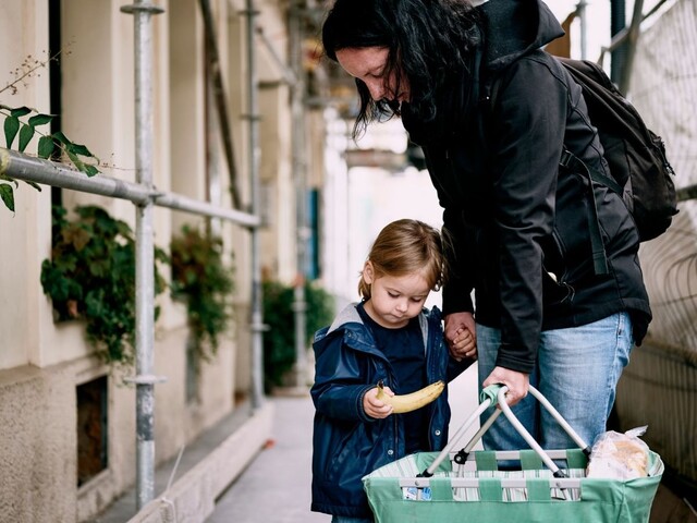 Eine erwachsene Person und ein kleines Kind stehen auf einem Gehweg mit Gerüststangen, halten gemeinsam einen grünen Einkaufskorb, und das Kind hält eine Banane in der Hand.