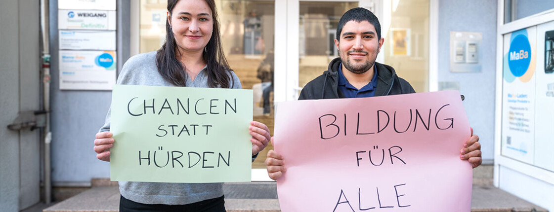 Zwei Personen halten Schilder mit „Chancen statt Hürden“ und „Bildung für alle“ vor dem Caritas-Bildungszentrum hoch