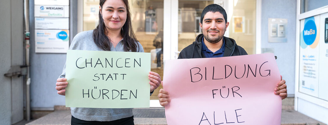 Zwei Personen halten Schilder mit „Chancen statt Hürden“ und „Bildung für alle“ vor dem Caritas-Bildungszentrum hoch