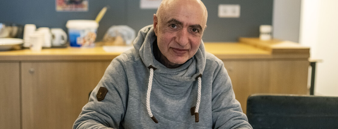 Ehemals obdachlose Person sitzt an Tisch und isst Suppe, im Hintergrund Küche mit Geschirr und Regalen