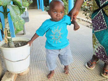 Kleines Kind in türkisfarbenem T-Shirt wird von einem Erwachsenen an der Hand gehalten und steht auf einem hellen Boden im Außenbereich.