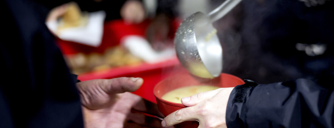 Eine rote Suppenschüssel wird mit einem Schöpflöffel mit dampfender Suppe befüllt. Eine Hand greift bereits zu Schüssel, um sie entgegenzunehmen.