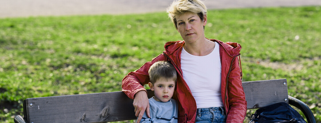 Eine erwachsene Person und ein Kind sitzen auf einer Parkbank in einer grünen Wiese, das Kind hält ein Stofftier, daneben steht eine Tasche.