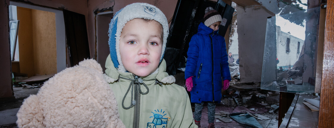 Ein kleiner Junge in Winterjacke und mit Haube hält einen Teddybären im Arm. Im Hintergrund sind die völlig zerstörten Möbel und Wände einer Wohnung zu sehen sowie auch ein traurig dreinschauendes kleines Mädchen.