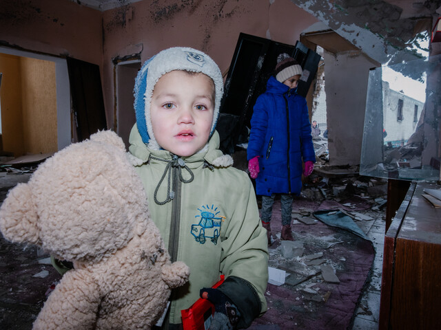 Ein kleiner Junge in Winterjacke und mit Haube hält einen Teddybären im Arm. Im Hintergrund sind die völlig zerstörten Möbel und Wände einer Wohnung zu sehen sowie auch ein traurig dreinschauendes kleines Mädchen.