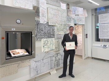 Künstler mit Tafel „Wiener städtische Versicherung“ in Raum mit Hängemaps und offenem Buch