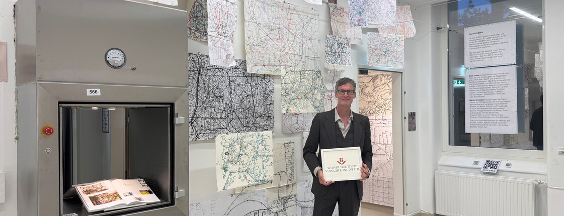 Künstler mit Tafel „Wiener städtische Versicherung“ in Raum mit Hängemaps und offenem Buch