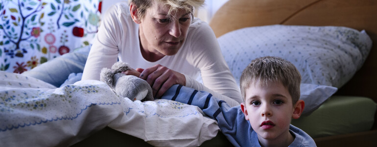 Eine erwachsene Person lehnt sich von einem Bett aus zu einem Kind, das davor steht, in einem Schlafzimmer mit Kissen, Decke und gemustertem Vorhang.