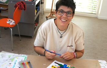 Künstler*in mit Brille malt mit Stiften ein farbiges Bild im Atelier der Caritas-Kunstgruppe im Weinviertel.