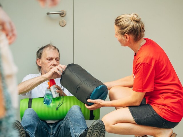Caritas Mitarbeiterin übergibt sitzendem Mann Schlafsack, Isomatte und Wasser vor einem Gebäude.