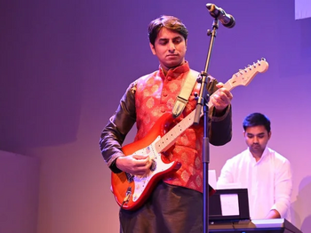 Zu sehen ist ein Gitarrist, der vor einem Mikrofon steht. Im Hintergrund spielt eine Person vermutlich Piano.