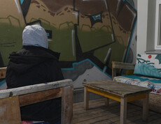 Jugendliche*r mit Kapuze sitzt allein auf Holzbank vor Graffitiwand im Außenbereich der Einrichtung