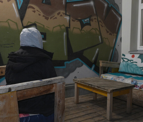 Jugendliche*r mit Kapuze sitzt allein auf Holzbank vor Graffitiwand im Außenbereich der Einrichtung