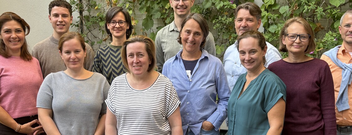 Das Team der PfarrCaritas steht auf einer Holzboden mit begrünter Mauer im Hintergrund
