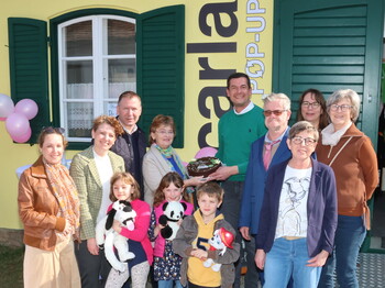 Eine Gruppe von Menschen steht vor einem gelben Gebäude mit grünen Fensterläden; einige Kinder halten Stofftiere, und in der Mitte wird eine dekorierte Torte präsentiert.