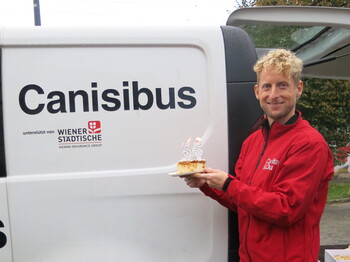 Mitarbeitender hält Kuchen mit Kerzen „35“ vor Canisibus-Fahrzeug der Caritas in Wien