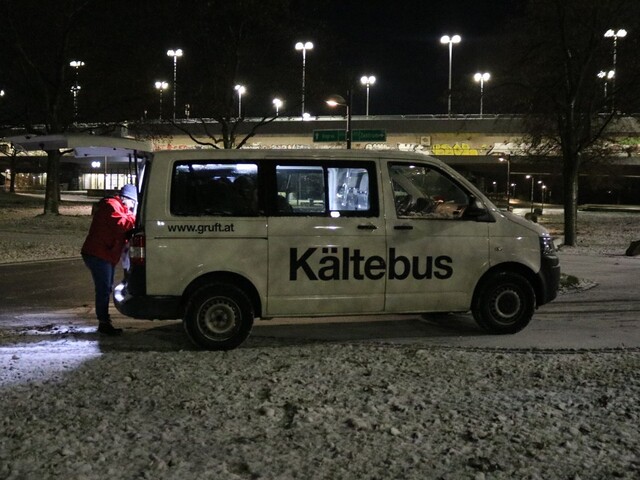 Der Kältebus der Gruft beziehungsweise Caritas steht vor einer Brücke in einer verschneiten Wiese. Eine Frau sucht etwas im Kofferraum des Busses.