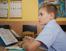 Ein kleiner junge sitzt an seinem Tisch in einem Klassenzimmer. Vor ihm steht ein offenes Lesebuch.