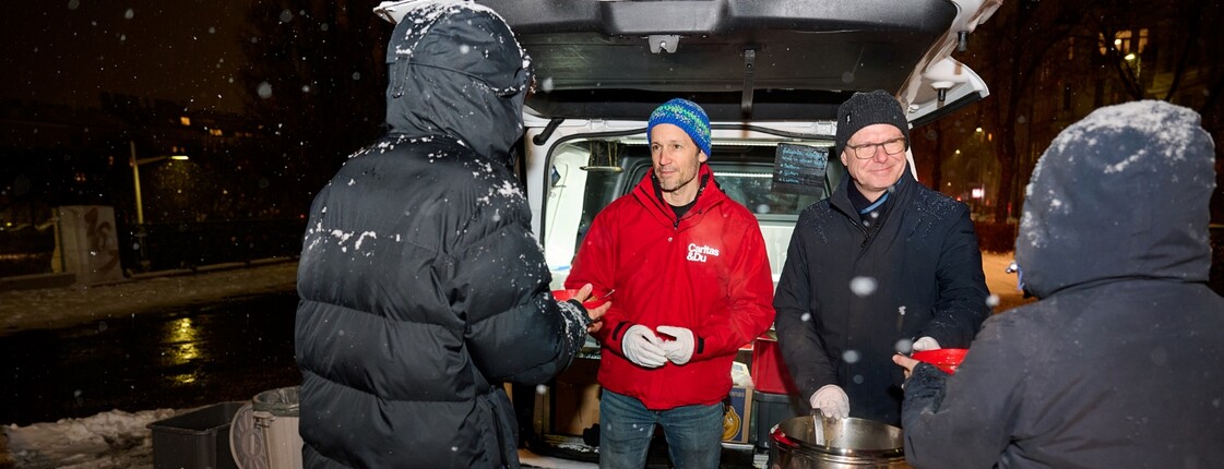 Zwei Caritas-Mitarbeiter geben beim Canisibus warme Suppe an obdachlose Menschen im Schneefall aus. © Drei-WirlPhoto