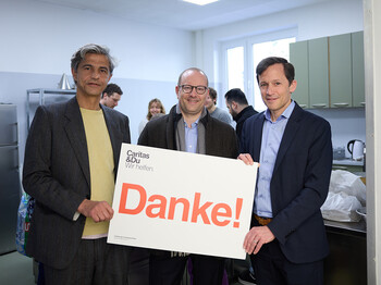 Drei Personen stehen in einem Küchenraum und halten gemeinsam ein Schild mit der Aufschrift „Danke!“ von Caritas & Du – Wir helfen.