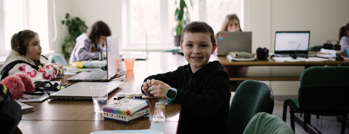 Ein Bub sitzt am Schreibtisch mit Unterlagen vor sich. Er lächelt in die Kamera. Im Zimmer sind im Hintergrund noch andere Kinder an den Schreibtischen und arbeiten.
