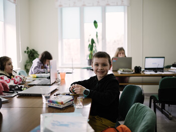 Ein Bub sitzt am Schreibtisch mit Unterlagen vor sich. Er lächelt in die Kamera. Im Zimmer sind im Hintergrund noch andere Kinder an den Schreibtischen und arbeiten.